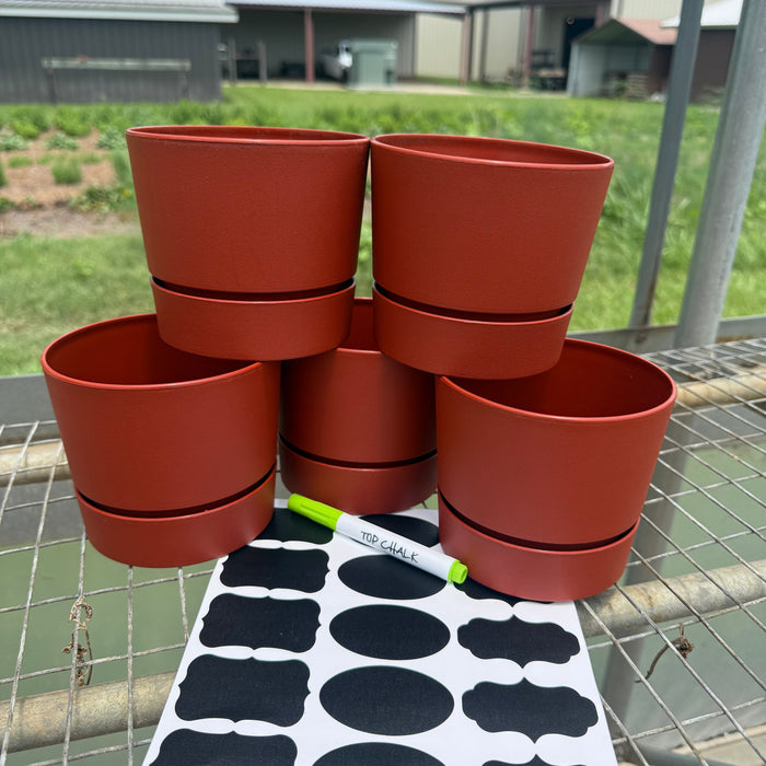 Five terracotta-colored plastic pot holders stacked on a wire table, with a sheet of black decorative labels and a green marker in front. An outdoor farm background with buildings and grass is visible behind the table.