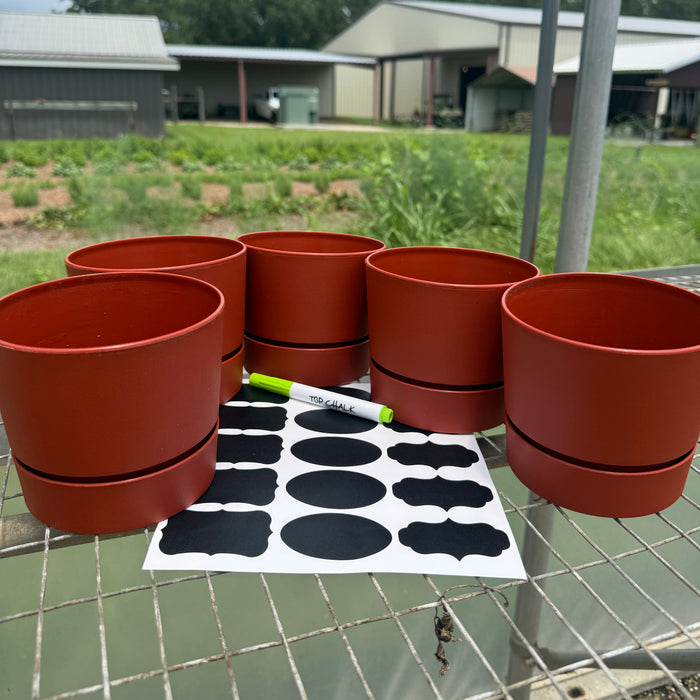 Five terracotta-colored plastic potting cups arranged on a metal table outdoors, with a sheet of black decorative labels and a green marker in front. A farm field and buildings are visible in the background.