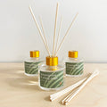 Three glass aroma diffusers with lemongrass and clary sage scent, featuring wooden reeds and gold caps, on a wooden surface.