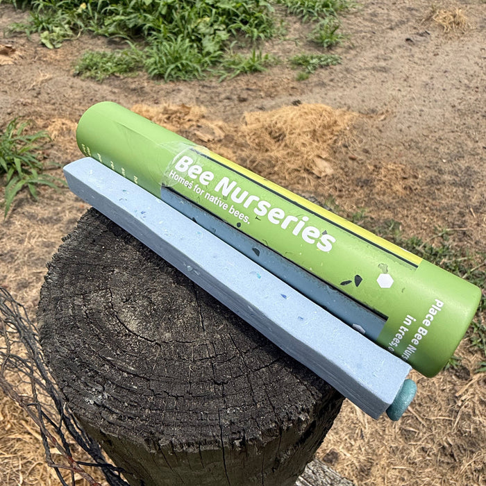 A green cylindrical beekeeping tool labeled 'Bee Nurseries' resting on a weathered wooden post in an outdoor garden setting.