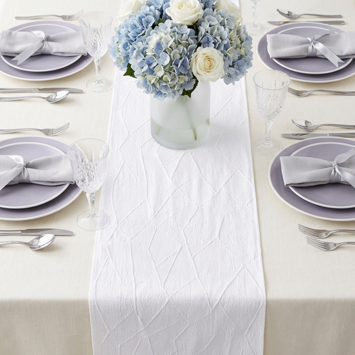 Embossed Geometric Jacquard Table Runner White on top an ivory tablecloth, gray charger plates, and gray/silver polyester napkins