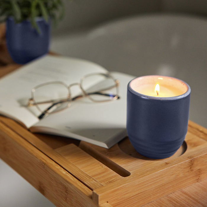 A blue ceramic candle with a lit wick sits on a bamboo bath tray next to a book and reading glasses.