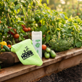 A gardening product set displayed on a wooden raised garden bed, featuring a bright lime green towel printed with the 'HOSS GARDENING CLUB' logo and a white canister of 'Greenhouse Cleaning Clay'. The background is a lush, out-of-focus garden with tomatoes, cucumbers, and green beans.