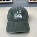 A washed dark green baseball cap with white embroidered text reading 'HOSS GARDENING CLUB' and a small plant graphic, displayed on a light countertop with a blurred store background.