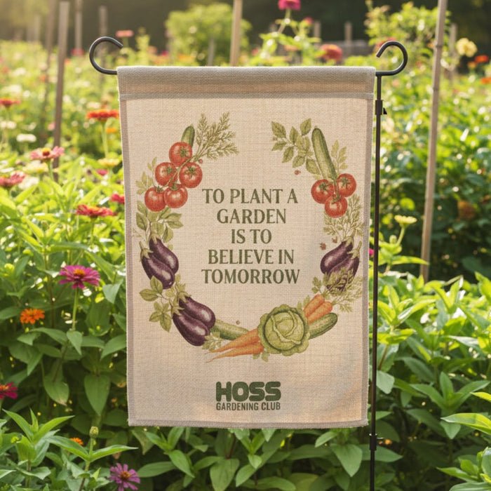 A rectangular garden flag hanging from a black metal stand in a sunny garden. The flag has a beige linen-style background with an illustrated wreath of vegetables and fruit around the words, 'TO PLANT A GARDEN IS TO BELIEVE IN TOMORROW.