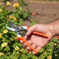 Hand holding orange-handled pruning shears with a garden background