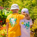 Two children standing outdoors in a garden, viewed from behind. The child on the left wears a bright yellow T-shirt with a large colorful floral bunny graphic and the words 'BUNN OF MARCH' on the back, along with a white bucket hat.