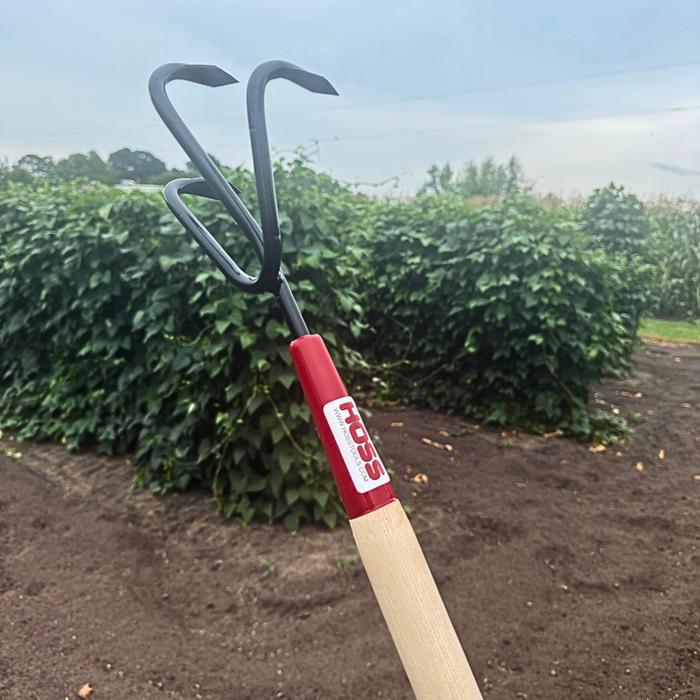 Triple Tine Cultivator with a red handle and visible brand logo, set against a background of green bushes and soil.